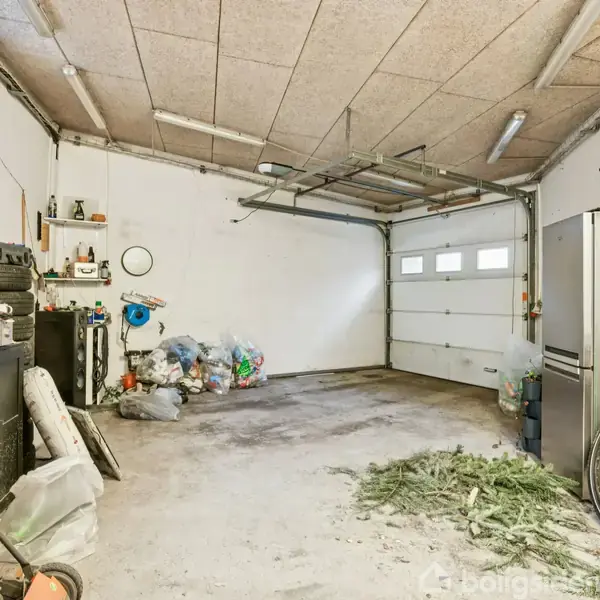 Garagen er rodet med poser og husholdningsartikler spredt rundt. En cykel står op ad højre væg ved siden af et køleskab. Der er en garageport bagerst.