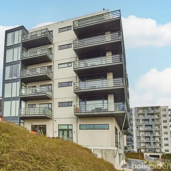 En moderne lejlighedsbygning med altaner strækker sig opad i et beboelsesområde. Ovenpå en forhøjning er bygningen omgivet af flere andre boligblokke og en klar, blå himmel.