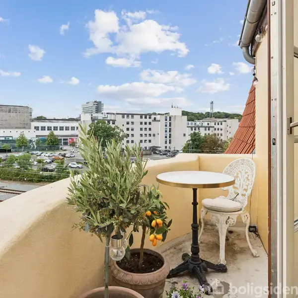 Terrassen har et lille bord og en stol placeret ved en halvmur. En plante står i en krukke. I baggrunden ses en byudsigt med bygninger og himmel.
