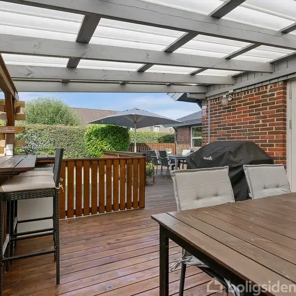 Overdækket terrasse med træbord og stole, grill med sort betræk, og parasol; omgivet af murstenshus og grønne hække i en baghave.
