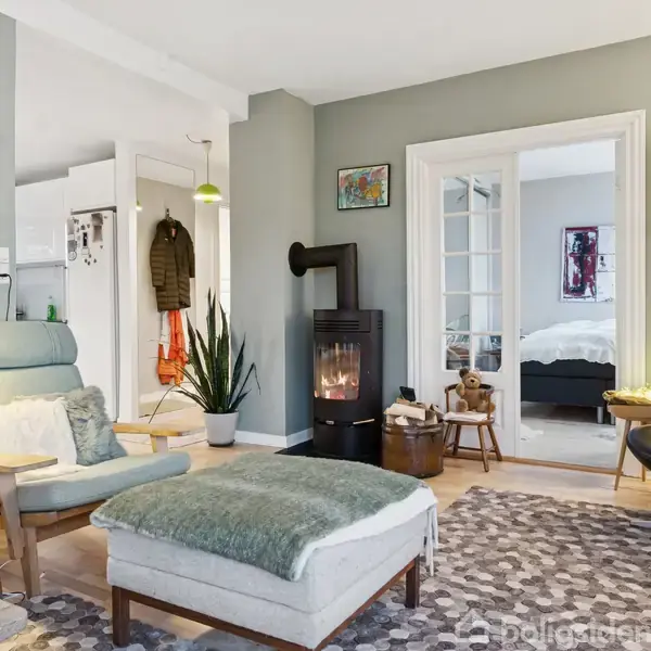 Lænestol med skammel står ved et sofabord i en stue med pejs. Rundt omkring er lamper, planter og billedkunst. Dør fører til soveværelse. Gulvtæppe dækker trægulv.