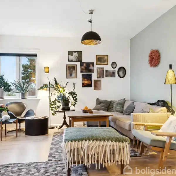 Stue med sofa og lænestol, dekoreret med tæppe og puder, omringet af planter og billeder på væggen; vinduer slipper naturligt lys ind, og der er moderne belysning.