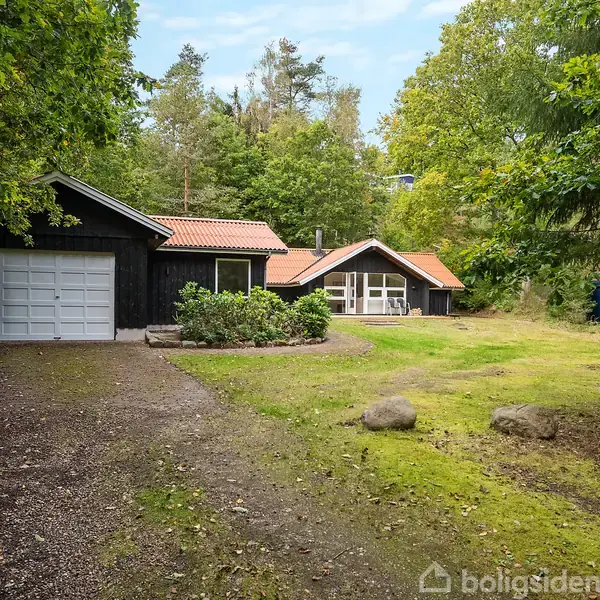 Sort hus med rødt tag står stille omgivet af grønne træer og en græsplæne. Der er en garage med hvid port til venstre og en indkørsel af grus foran.