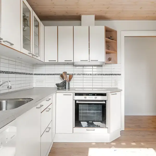 Et moderne køkken med hvide skabe og skuffer, en indbygget ovn på en trægulvsoverflade. Vask og hane er placeret til venstre, omkranset af hvide flisede vægge.