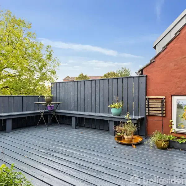 En udendørs terrasse med grå træplanker har en blåmalet bænk. Der er flere planter i krukker. Rød murstensvæg med et airconditionanlæg. Træer og blå himmel ses i baggrunden.