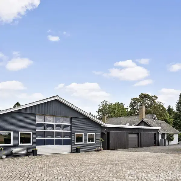 En grå bygning med store vinduer står stille. Omgivet af en brostensbelagt gård, ses træer og en blå himmel med skyer.