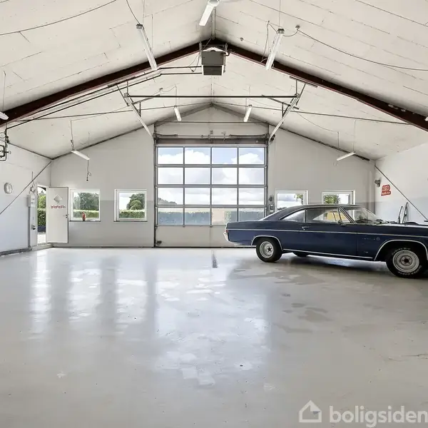 En tom garage med en blå klassisk bil parkeret. Der er store vinduer og højt til loftet, hvilket giver et lyst rum. Der er ingen synlig tekst.