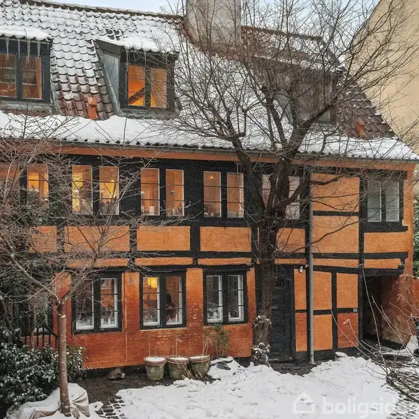 Et historisk bindingsværkshus med sneklædt tag, står stille i en vinterhave. Omkring huset ses træer uden blade, en snedækket gårdhave og flere vinduer med lys indefra.