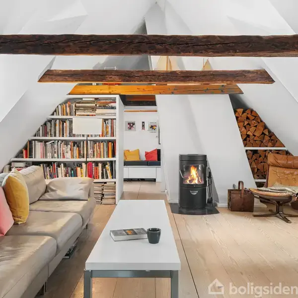 To barstole står ved en køkkenø, hvor køkkenredskaber hænger. Bagved er et køkken med grå skabe, store vinduer og en trædetaljeret gulv. Et dekorativt skat på væggen.