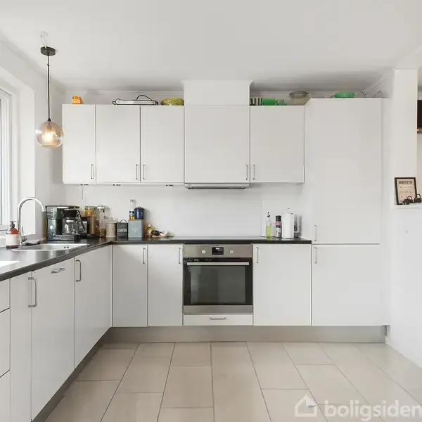 Et hvidt køkken med garderober og skuffer. En indbygget ovn i skabsvæggen. Flere køkkenapparater står på bordpladen under et vindue. To lamper hænger fra loftet.