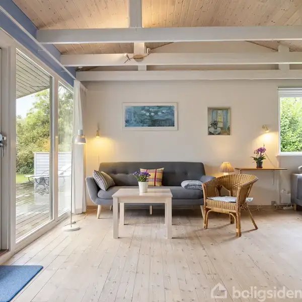 Stue med gråt sofa-arrangement og træstol ved lavt bord, udsigt til terrasse gennem store vinduer. Vægkunst og lamper tilføjer hygge; pejs ses i det lyse interiør.