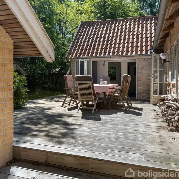 En træterrasse har et bord med en dug og seks stole rundt om det. Terrassehuset er omgivet af murstensvægge og grønne omgivelser med sollys, der filtrerer gennem træerne.