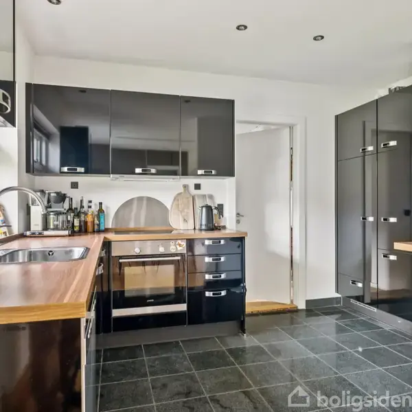 Køkken med sort-skabsindretning, træbordplader, og ståludstyr. En ovn og vask er tydelige. To barstole står ved en lille disk. Lyse vægge og mørkt klinkegulv ses.