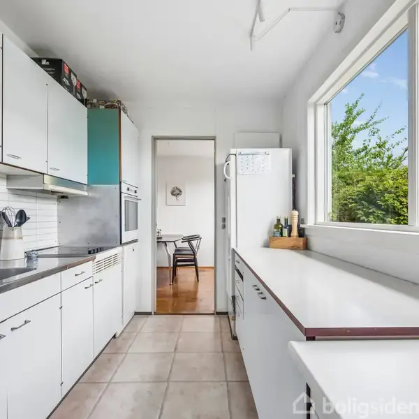 Køkken med hvide skabe og stålbordplade, hvor der står nogle køkkenredskaber. Store vinduer lader masser af lys ind. I baggrunden ses en åben dør mod en spisestue.