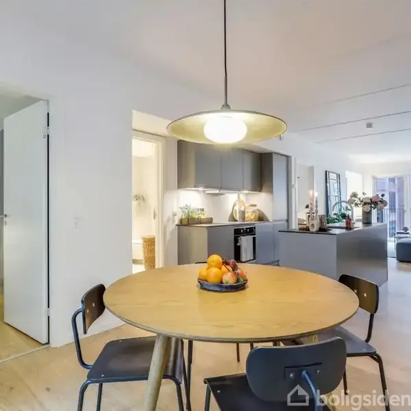 Rundt spisebord med frugtfad står i et moderne, åbent køkken-alrum. Bagved ses køkkenet med grå skabe og stue med sofagruppe. Et stort vindue giver lys til rummet.