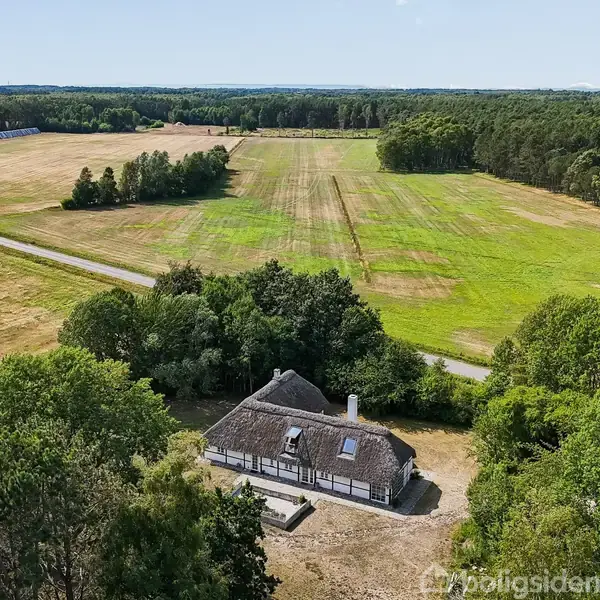 Stråtækt hus omgivet af frodige træer; placeret ved en stor, åben mark ved siden af en vej. Landskabet er domineret af grønne marker og skovområder i baggrunden.