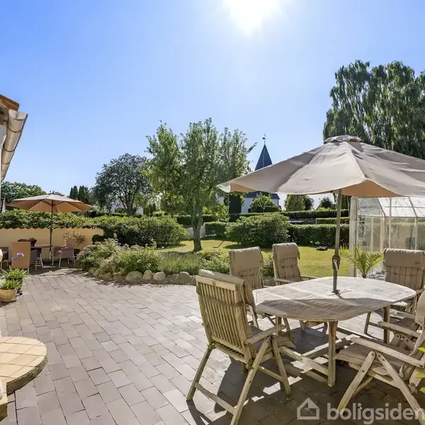 Terrassemøbler med parasoller står på en flisebelagt terrasse i en have. Omkring er der græsplæne, træer og en kirketårn i baggrunden under en klar blå himmel.