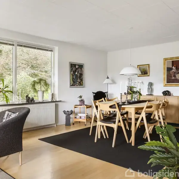En spisestue med et træbord og seks stole på et sort tæppe. Væggene har kunstmalerier, og store vinduer giver udsigt til naturen udenfor. Planter pryder rummet.