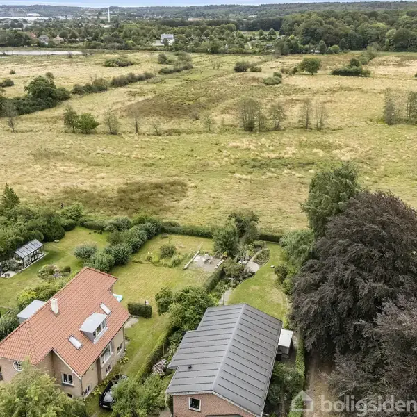 To huse med røde og grå tage i en frodig have omgivet af træer, med en stor grønt område og bakker i baggrunden. Vandoverflade kan ses i horisonten.