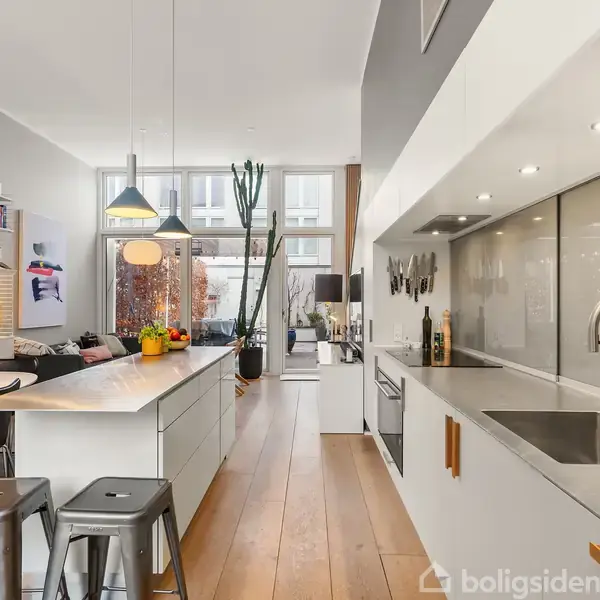 Et moderne køkken med en stor, hvid køkkenø og høje stole, omgivet af åben stue med sofa, hylder og store vinduer. Belysning hænger over køkkenøen. Ingen tekst.