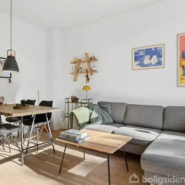 Grå sofa med puder står i hjørnet af en stue ved siden af et træbord med sorte stole. Væggene har plakater, og en lampe hænger over bordet.