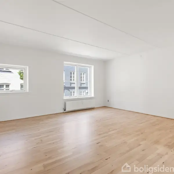 Et lyst rum med trægulv og hvide vægge er tomt. To vinduer lader lys strømme ind, og udenfor ses en bygning. Ingen tekst er til stede.