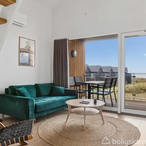 En stue har en grøn sofa og en rund tæppe foran et sofabord. Store glasdøre åbner til en terrasse med udendørsmøbler og udsigt over et landskab med huse i baggrunden.