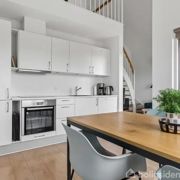 Køkken med hvidt skab og integreret ovn, kaffemaskine på bordet. Spiseborde har grøn plante i kurv. Trappe bagved leder til anden etage, vindue på højre side.