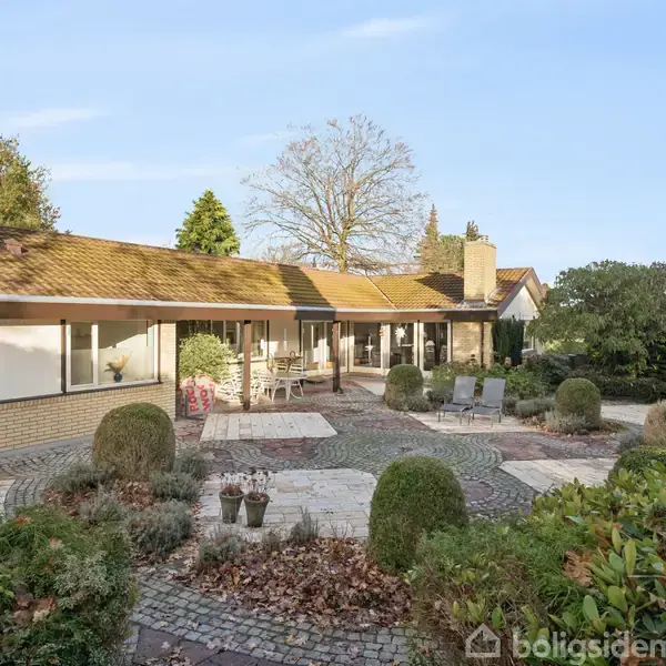 En étplans-villa med store vinduer og gule mure står omringet af en velplejet have med flisebelagt terrasse og buske, mens sirlige stier fører gennem det grønne område.