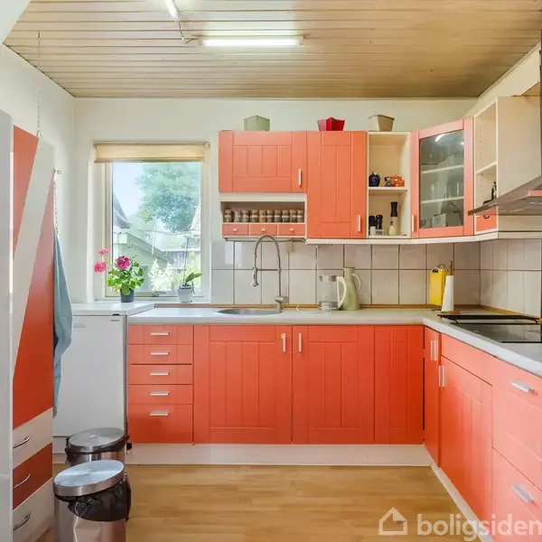 Et køkken med orange skabe og hvid bordplade, står tomt. Rummet er lyst med et vindue, komfur, vask, og køleskab, omgivet af blomster og køkkenredskaber.