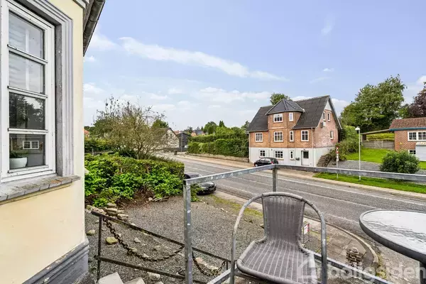 En tom stol står på en terrasse ved siden af et rundt bord. I baggrunden ses en vej og et murstenshus omringet af grønt i et boligområde.