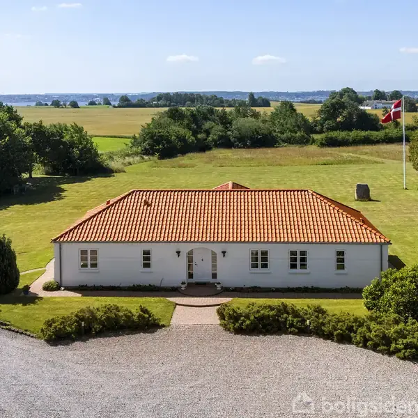 Hvidt hus med rødt tegltag står i en grøn have. En dansk flagstang står til højre. Grusbelagt indkørsel fører til huset. Skov og marker er i baggrunden.