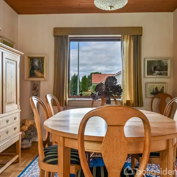 Spisebord med seks stole står i en spisestue. Store vinduer viser udsigt til en have. Væggene er dekoreret med malerier, og der er et skab til opbevaring.