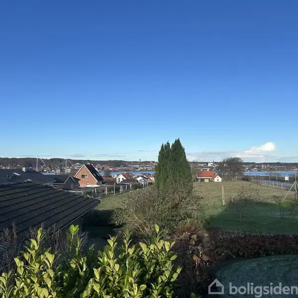 Et landskab med en række huse i forgrunden. Baghaven og et træ er synligt. I baggrunden ses en fjord og blå himmel.
