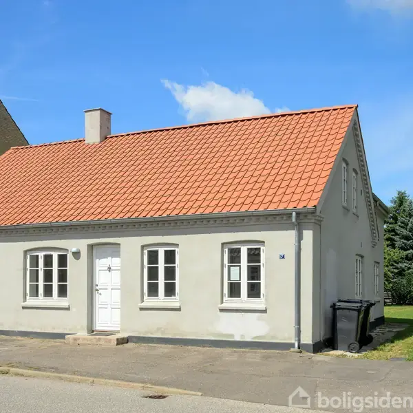 Hus med hvid facade og rødt tegltag står roligt ved siden af en vej. To sorte beholdere ses ved siden af huset, med græs og træer i baggrunden.