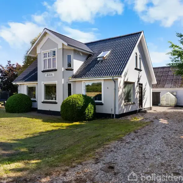 Et hvidt hus med sort tag står på en velplejet græsplæne omgivet af buske, træer og en grusbelagt indkørsel under en blå himmel.