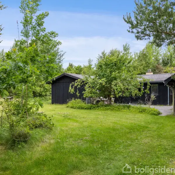Sort træhus står stille med terrassebord og stole i en flisebelagt gårdhave. Omkranset af frodig grøn bevoksning, skaber det en fredelig atmosfære.