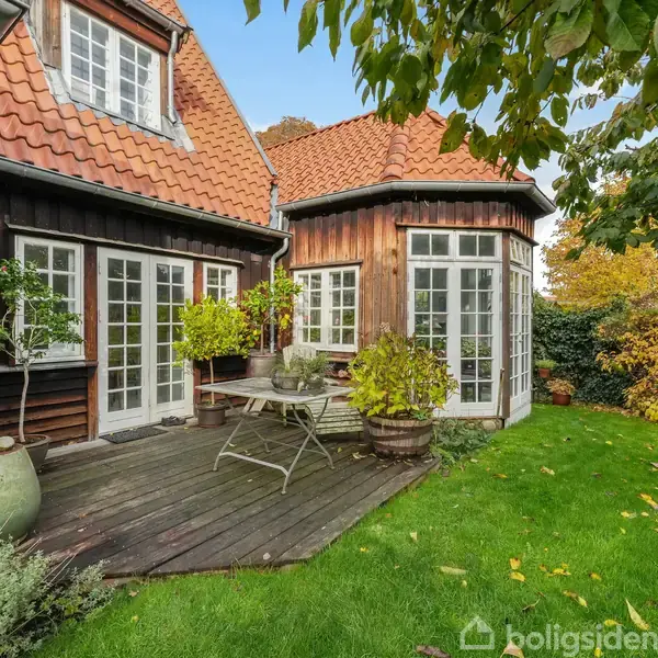 Træhus med rødt tegltag har en træterrasse med bord og planter. Terrassen omgiven af velplejet græs og buske under en blå himmel.