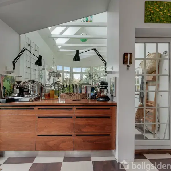 Et køkken med træskuffer står under to væglamper ved et stort spejl. En dør til højre åbner op mod en møbleret veranda med glasvægge. Skakternet gulv.