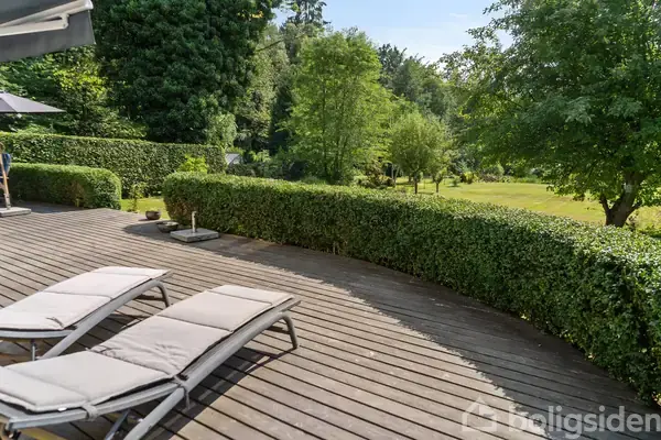 To liggestole står på en træterrasse, omgivet af en velplejet hæk. I baggrunden ses en stor have med træer og grønne områder.