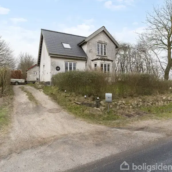 Hus står stille i en landlig kontekst omgivet af træer og buske. En grussti fører op til forhaven og indgangen, med en bil parkeret i baggrunden.