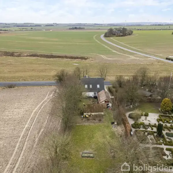 Et hus med tilhørende bygninger ligger midt i et landbrugsområde. Bag huset strækker marker sig ud i det fjerne, og en vej løber langs med markerne. En velholdt have er til højre.