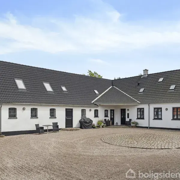 En hvid, L-formet bondegård med sort tegltag står på en grusbelagt gårdsplads, omgivet af træer. Flere vinduer og døre ses langs bygningens facade.