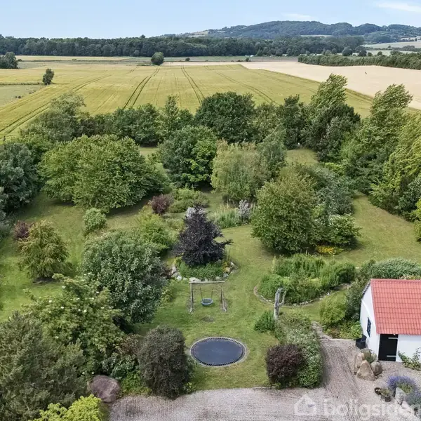 En stor have med træer og en trampolin i midten omgivet af grønne marker og bakker. Et hvidt hus med rødt tag ses i højre hjørne.