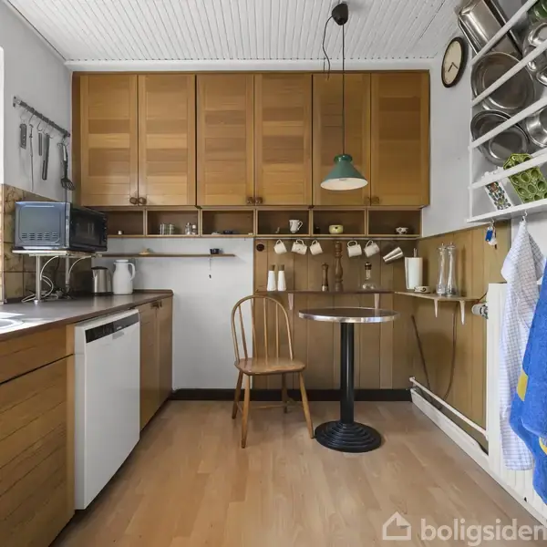 Et træbord med en stol står i et køkken med træskabe og en væghylde fyldt med tallerkener og kopper. En lampe hænger over bordet, og der er en blå viskestykke.