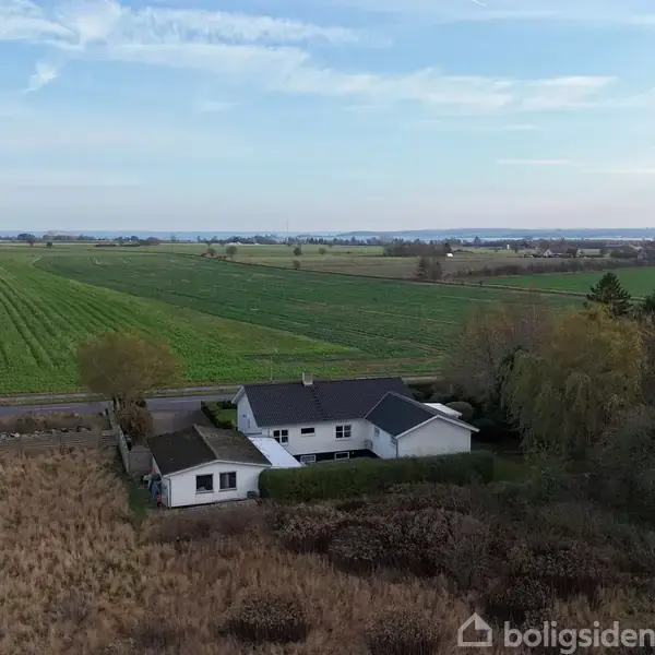 En hvid villa med sort tag ligger stille omgivet af grønne marker og spredte træer. I baggrunden er der skov og klar blå himmel, der skaber en fredelig landlig atmosfære.