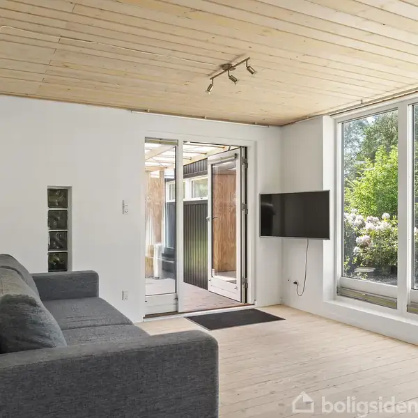 En grå sofa står i en moderne stue med trægulve og lyst loft. Vægmonteret fladskærms-TV overfor sofaen. Store vinduer giver udsigt til en grøn have.