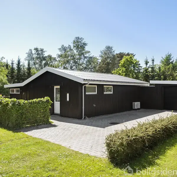 Et sort træhus står stille omgivet af en grøn have og hække. En flisebelagt indkørsel fører op til huset, beliggende i et skovrigt område under en klar blå himmel.