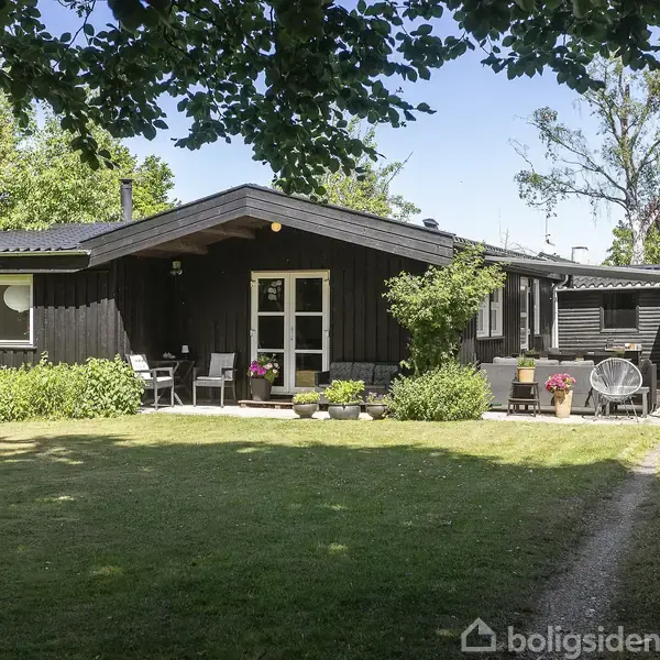 Et sort træhus med store vinduer, omgivet af en velplejet have med græsplæne og blomsterbede. Havemøbler under overdækket terrasse skaber en hyggelig stemning i solrigt vejr.