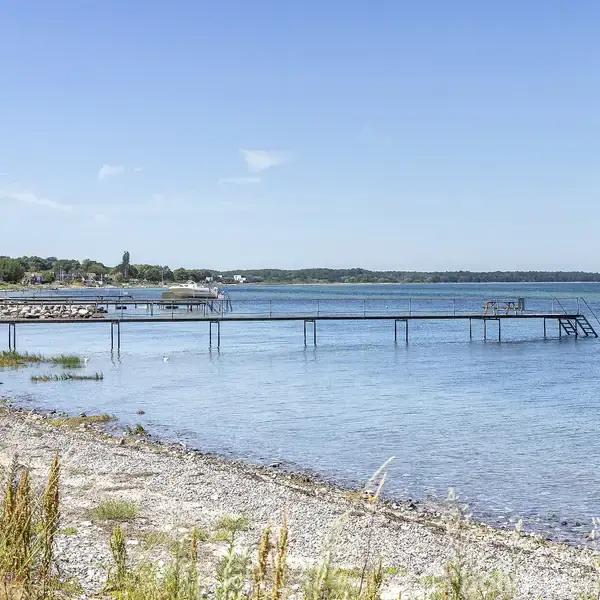 En lang badebro strækker sig ud i det blå, rolige hav fra en stenstrand, omgivet af sparse vegetation. Fjern beboelse og træer markerer horisonten under en klar blå himmel.